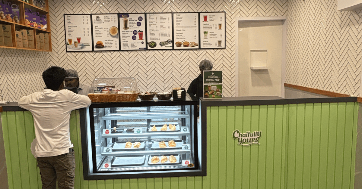 Interior of Chai Kings Ameerpet outlet featuring a green front counter with a pastry display, menu boards on a white patterned wall, warm lighting, and a customer ordering at the counter.